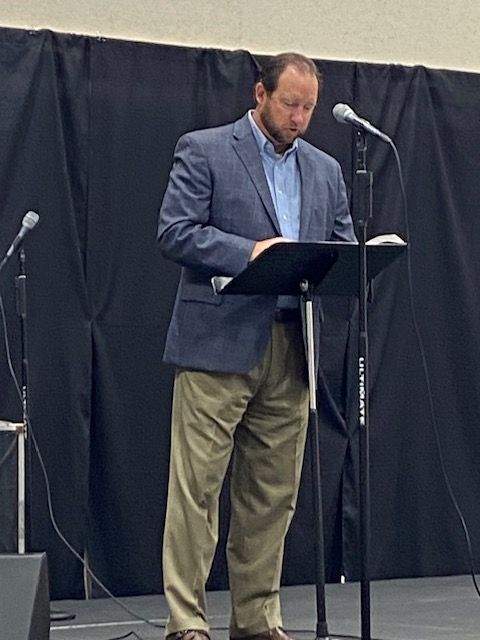 A man is standing at a podium reading a book and singing into a microphone.