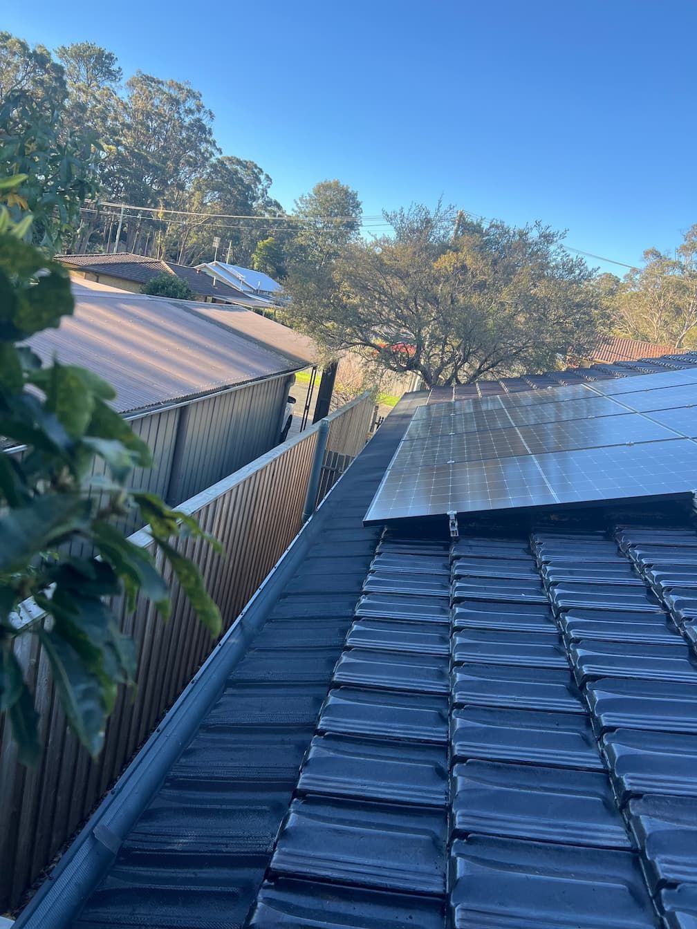 Rooftop with solar panels and dark tiles next to a fence and trees on a sunny day.