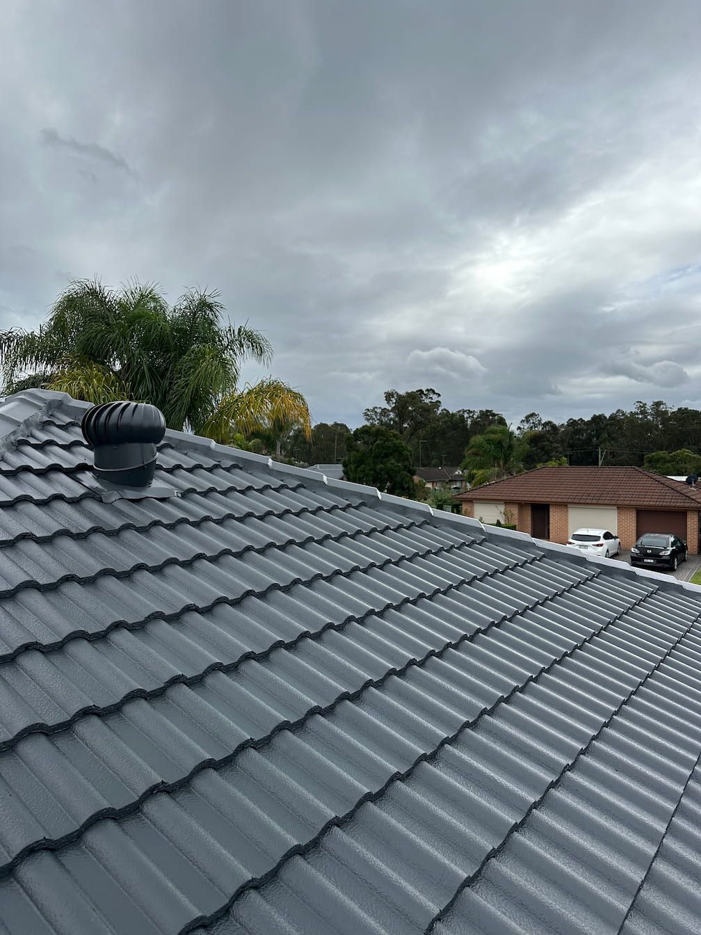Gray tiled roof with a vent, against a cloudy sky, suburban homes visible in the distance.