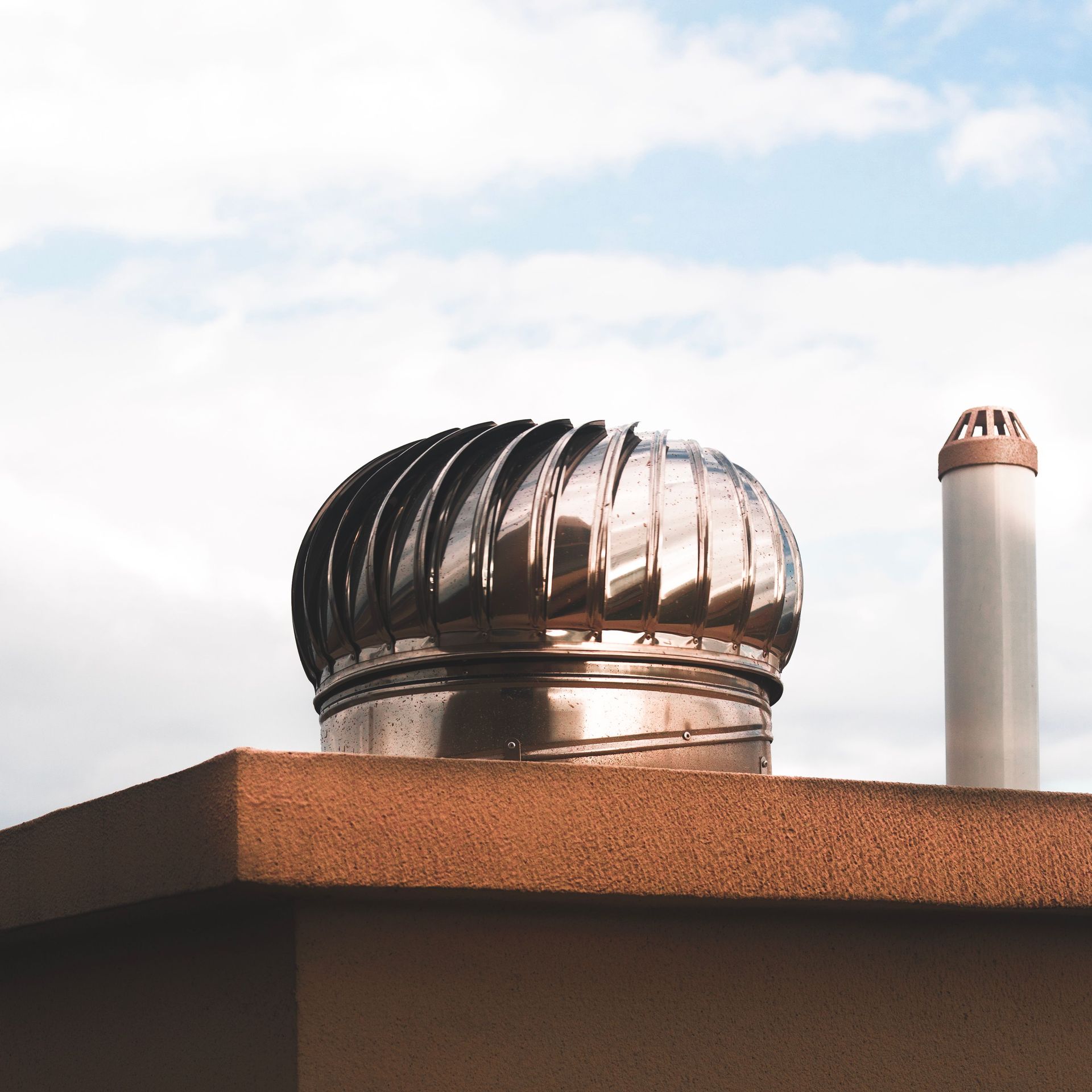 Metal rooftop vent with spinning turbine and a white chimney against a cloudy sky