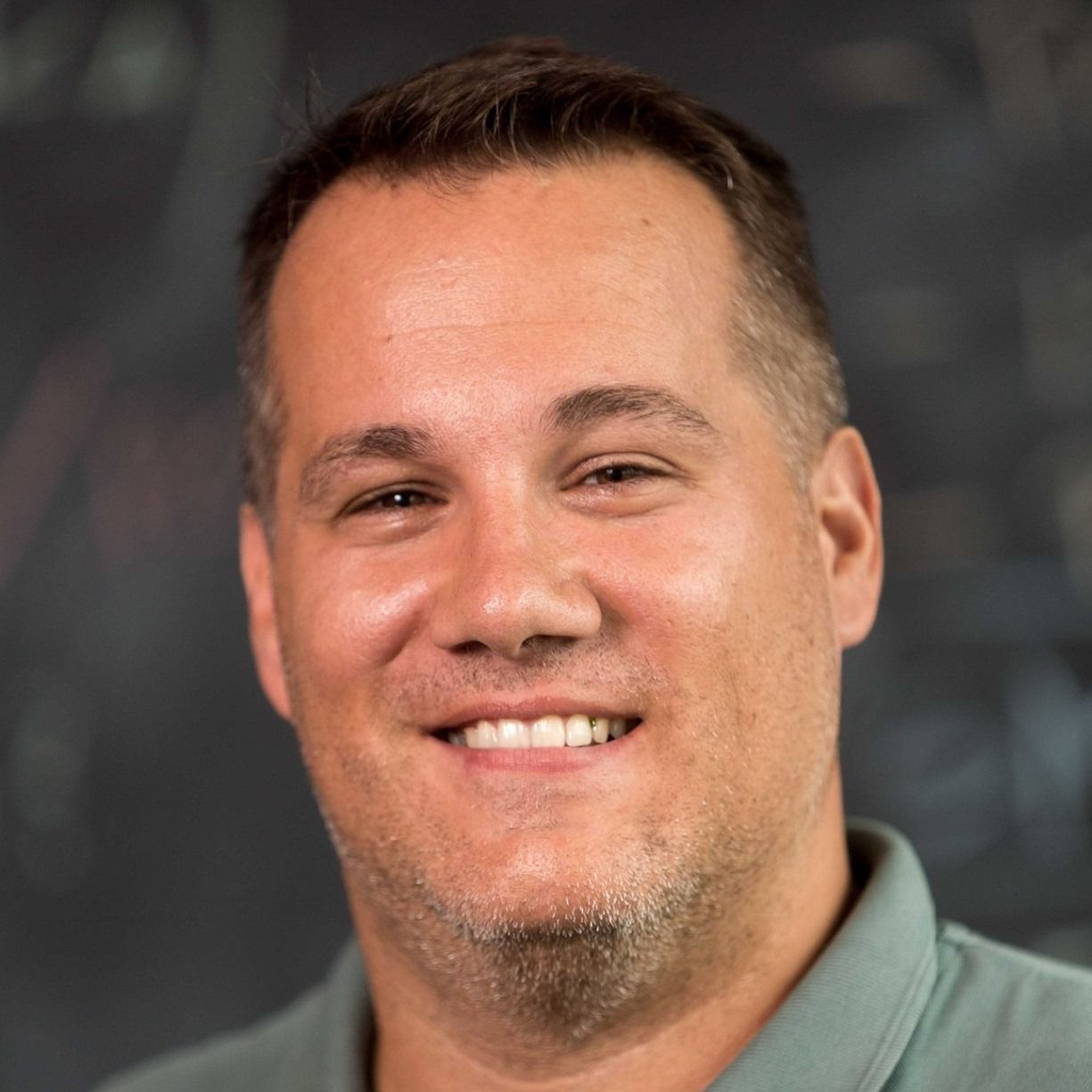 Man with short brown hair smiling, wearing a teal polo shirt. Dark background.