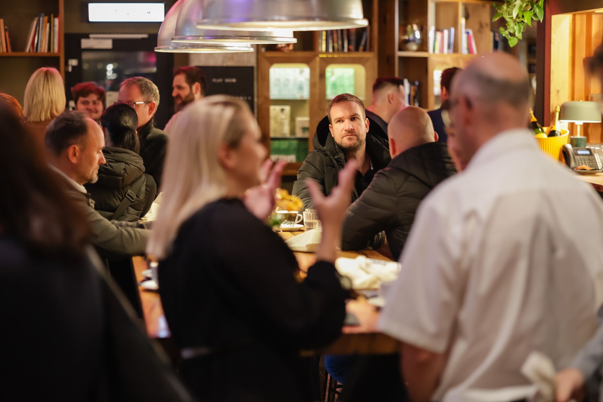 Ljudje se zbirajo v baru. Ženska gestikulira med govorjenjem. Drugi gostje sedijo za barom in v ozadju.
