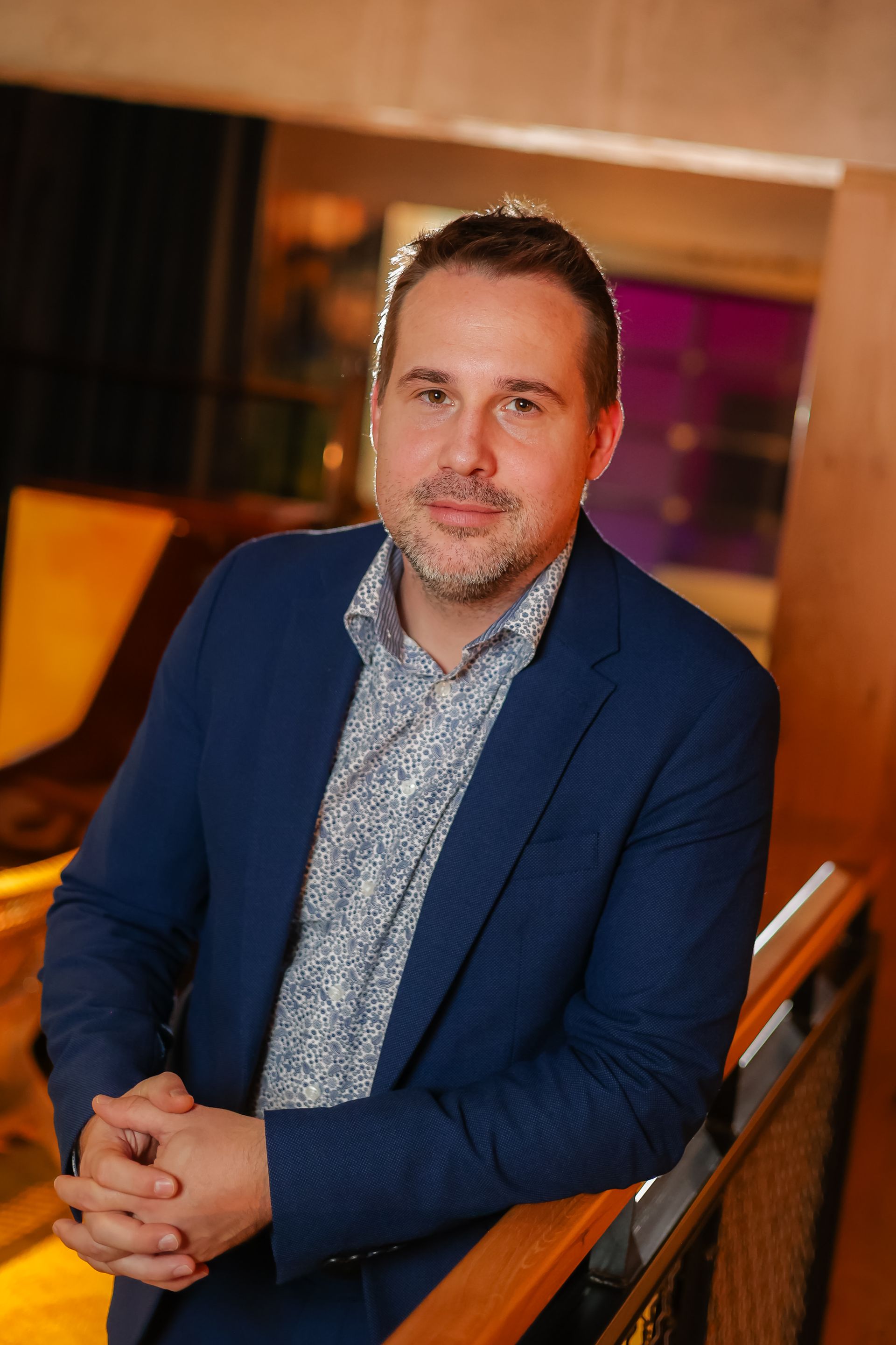 Man in a blue blazer leans on a railing, smiling. He wears a patterned shirt in a dimly lit space.