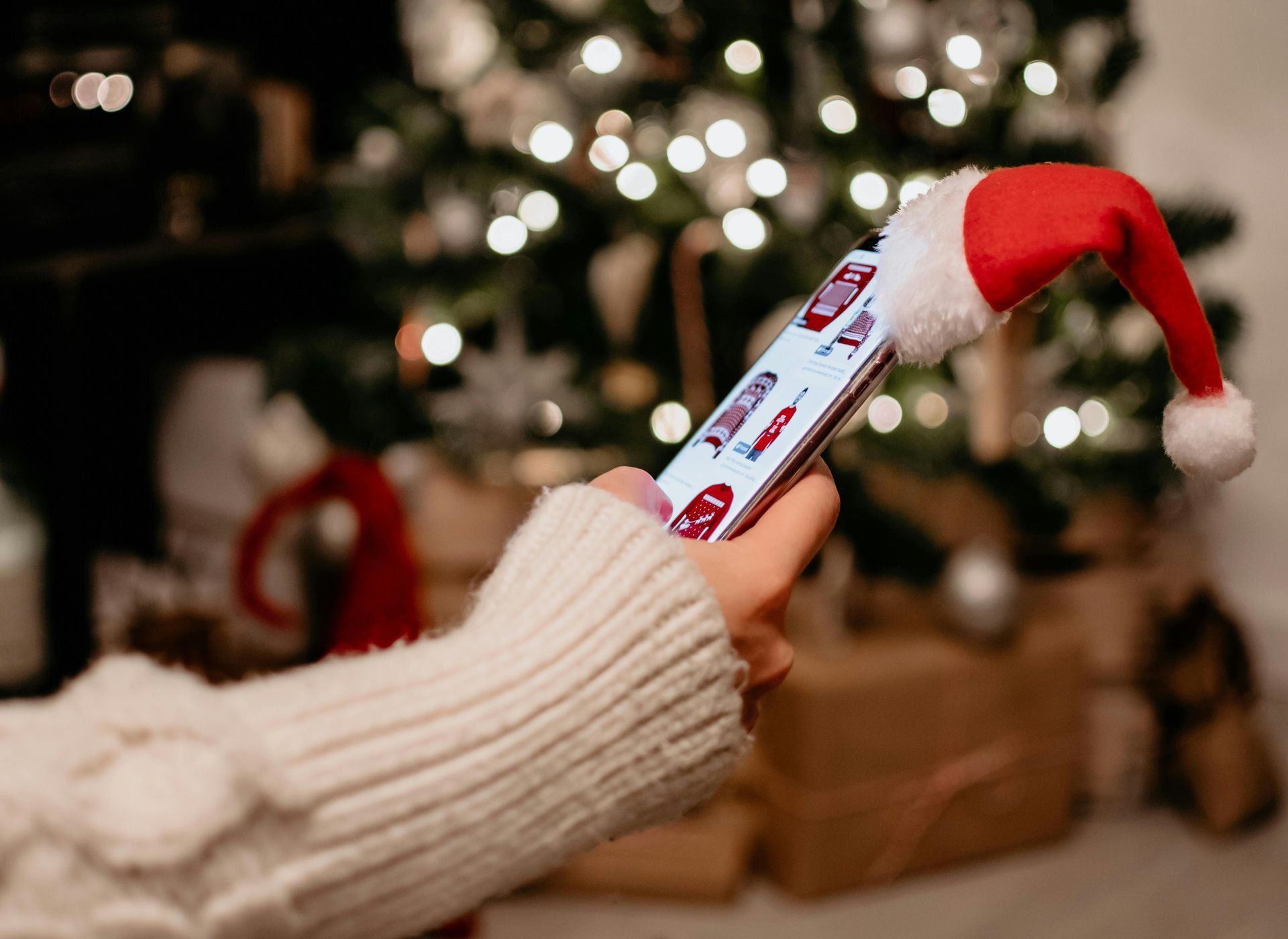 Hand holding a smartphone with a Santa hat, shopping online with a decorated Christmas tree in back.