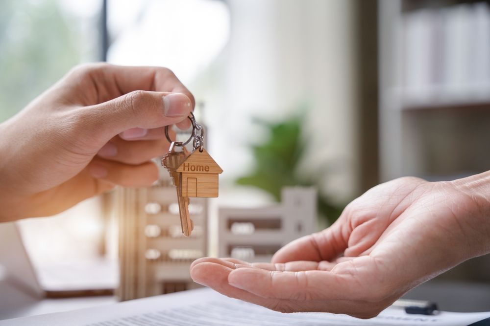 A hand hands a set of keys with a house-shaped keychain to another hand, suggesting a real estate or home-buying deal.
