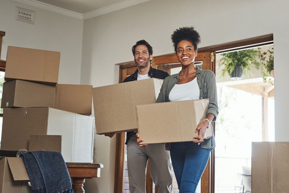 A couple smiling while carrying cardboard moving boxes into a bright, unfurnished room.