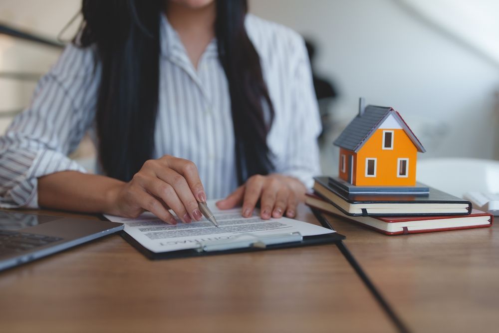 A person in a striped shirt signs a document near a small model house sitting on a stack of notebooks on a wooden desk.