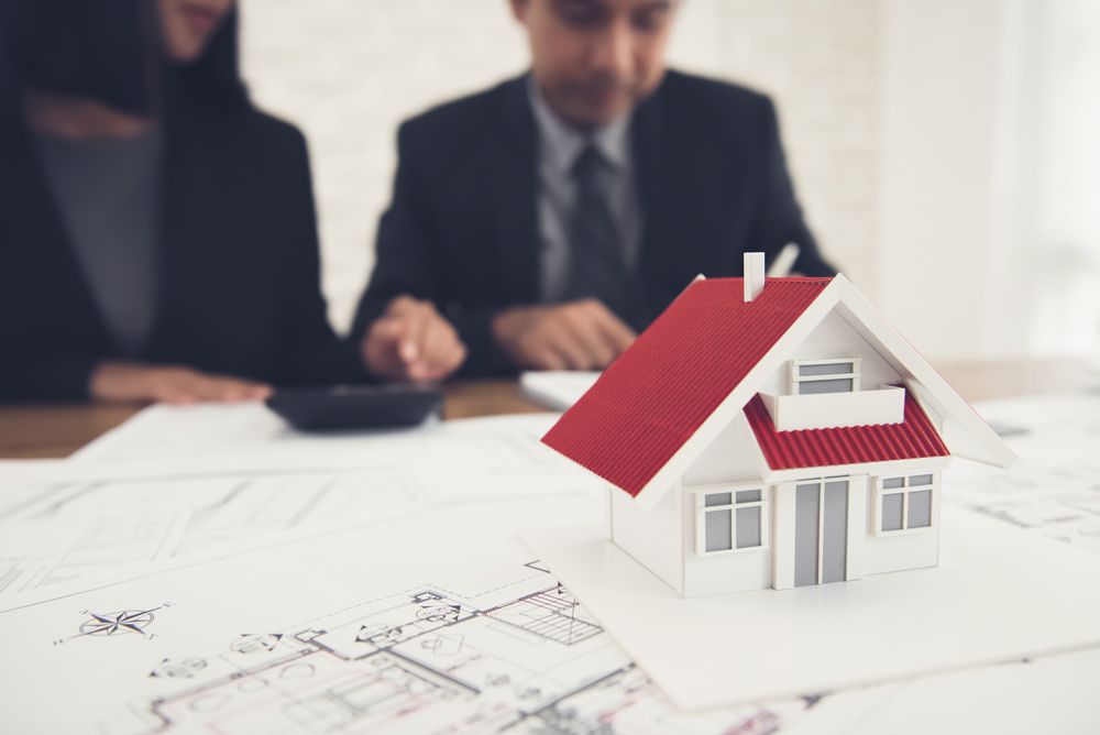 A small scale model house with a red roof sits on architectural floor plans in front of professionals in suits.