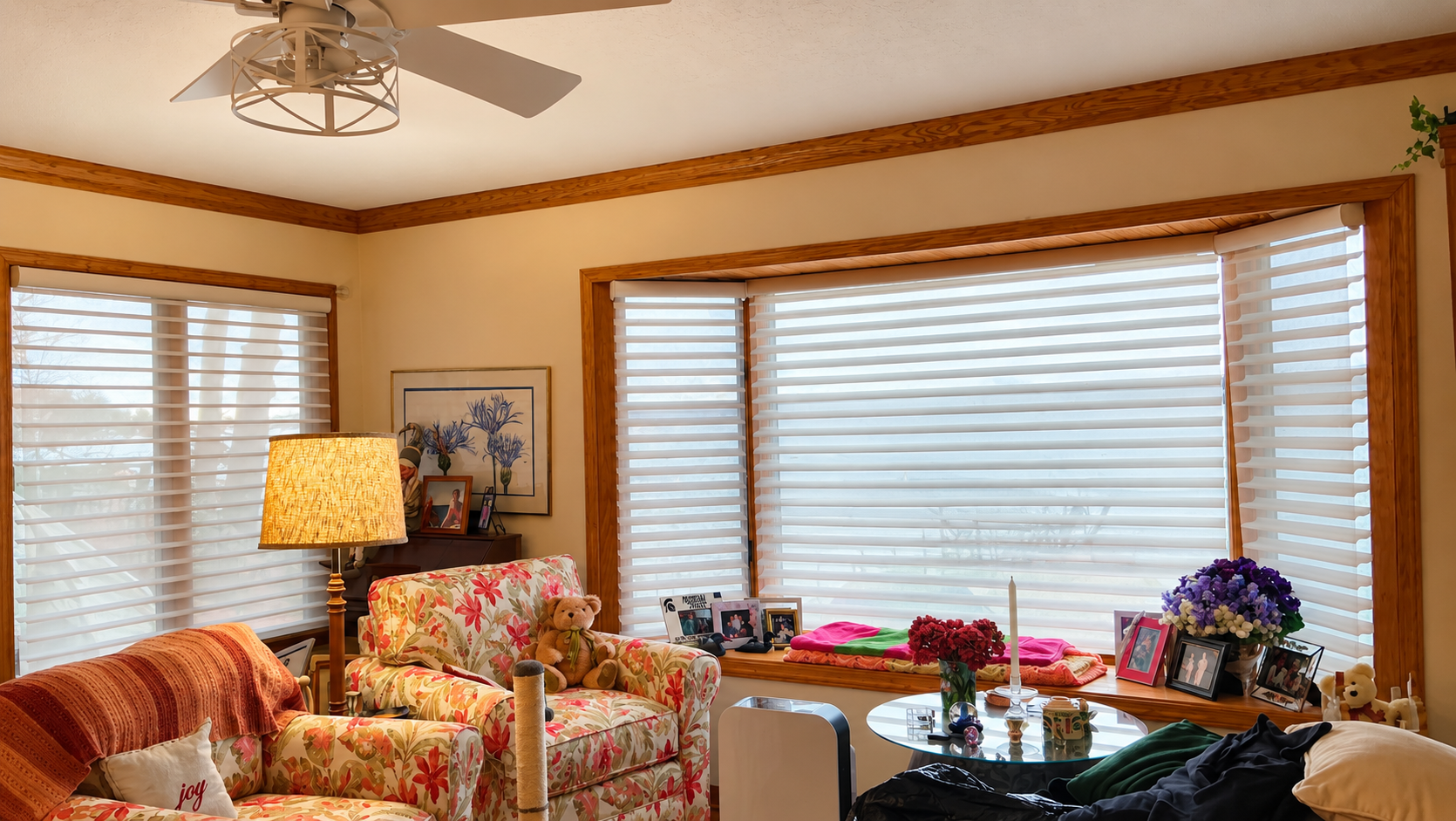 Bright living room with floral chairs, large windows with blinds, and a coffee table with flowers.