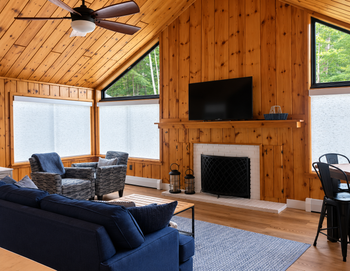 Cozy wood-paneled living room with sofa, chairs, fireplace, TV, and large windows