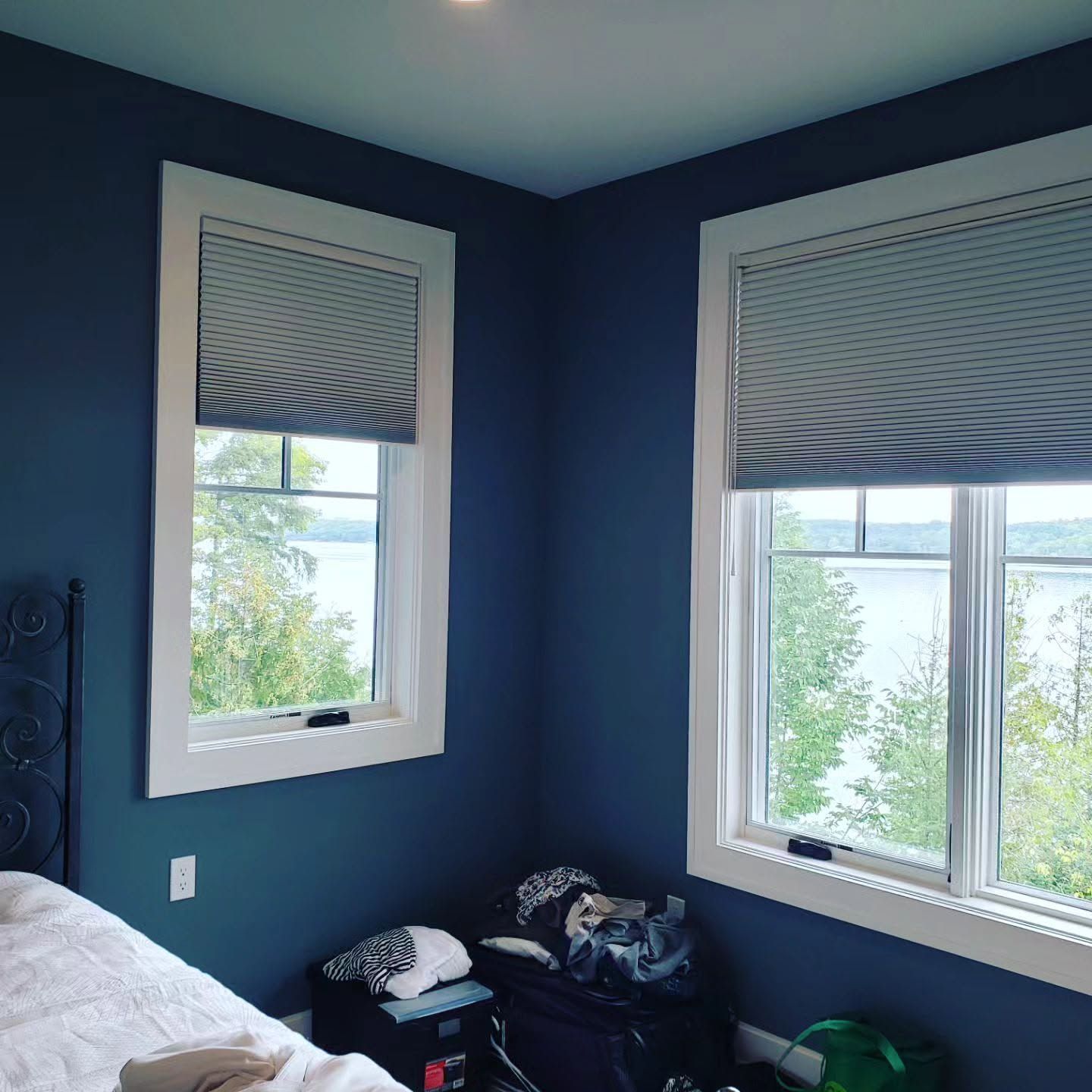 Blue bedroom corner with two windows, white blinds, and clothes piled near a bed.