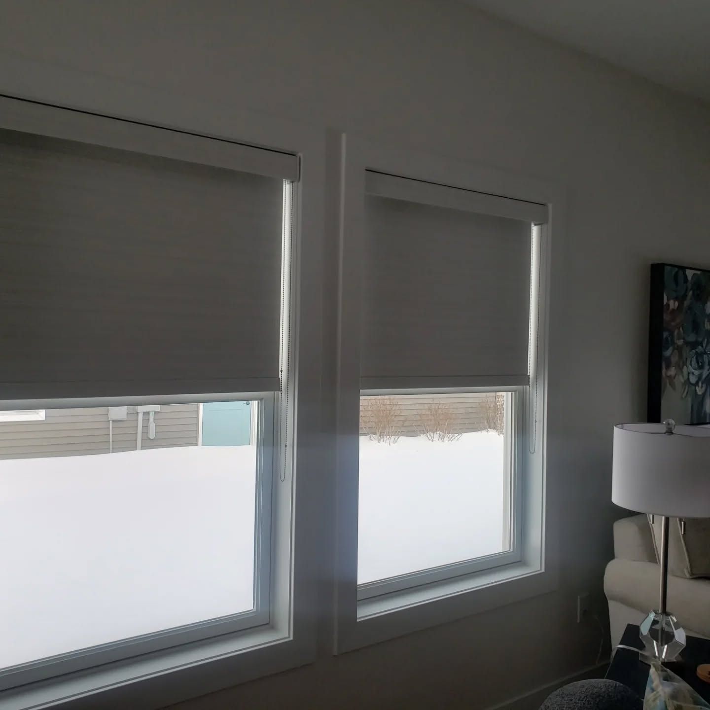 Two tall windows with white blinds in a bright room, with a lamp and sofa at the right edge.