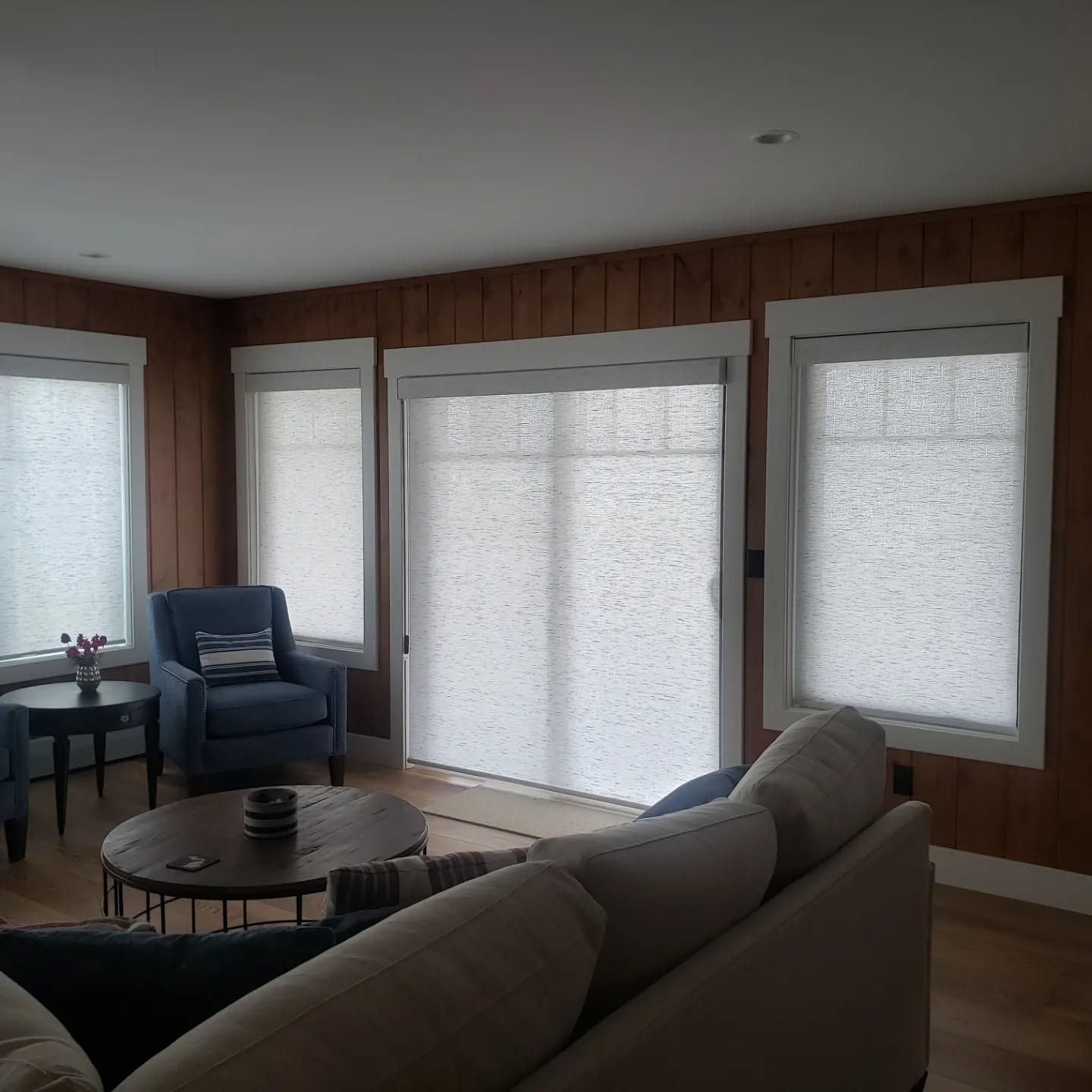 Living room with large windows covered by white shades, blue chair, coffee table, and gray sofa