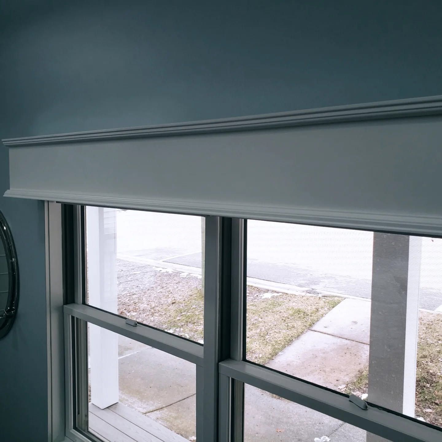 Window with white blind partially raised, showing a bright outdoor sidewalk and grass outside.