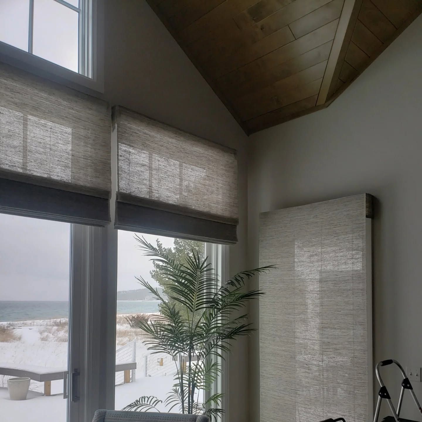 Bright room with large windows, woven shades, a potted palm, and a snowy beach view outside.