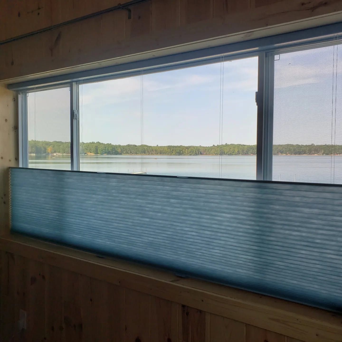 Blue window shade inside a wooden room, looking out over a calm lake and distant tree-lined shore