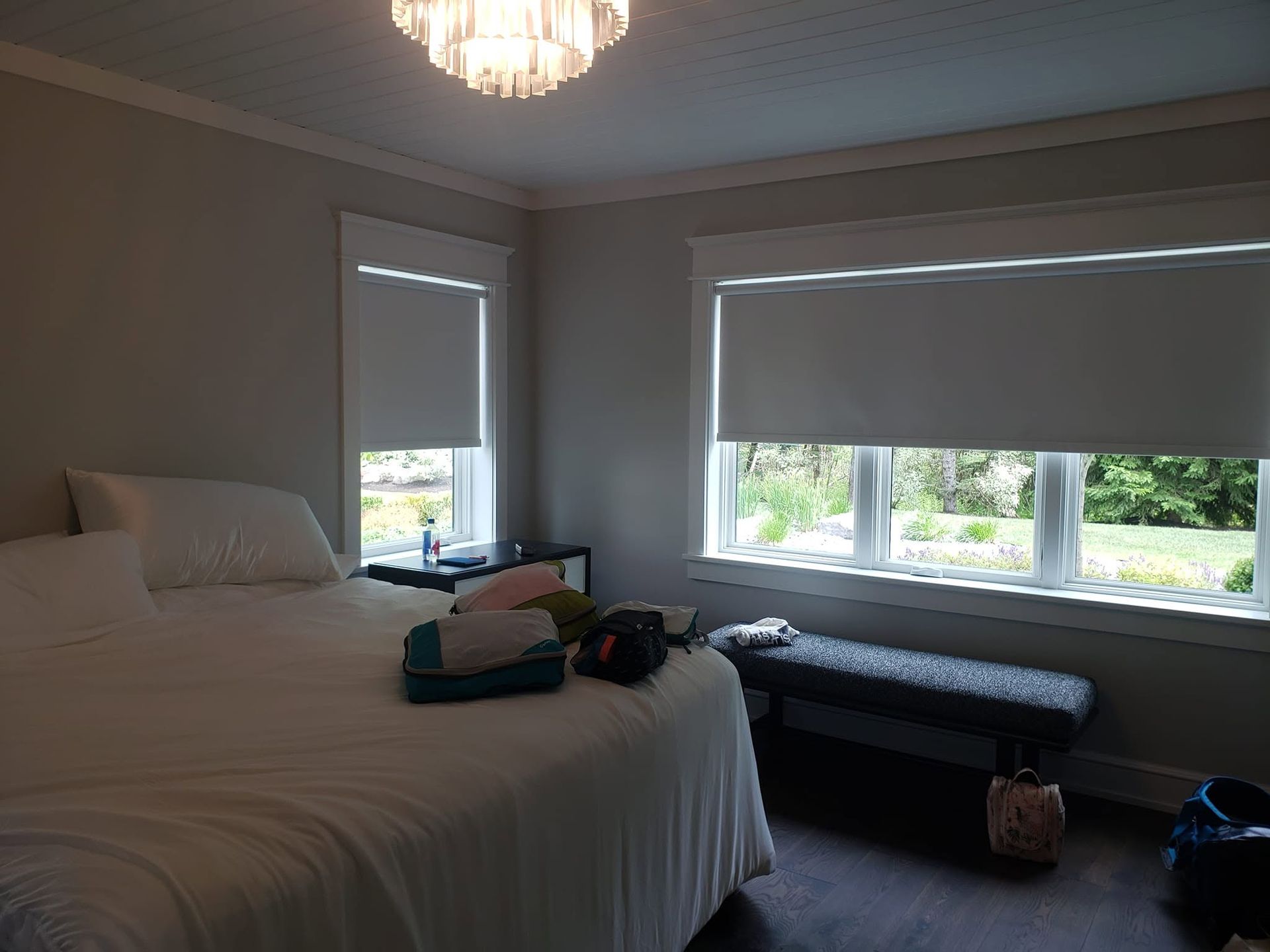 Bedroom with a made bed, bench, and two windows with blinds, lit by daylight and a ceiling light