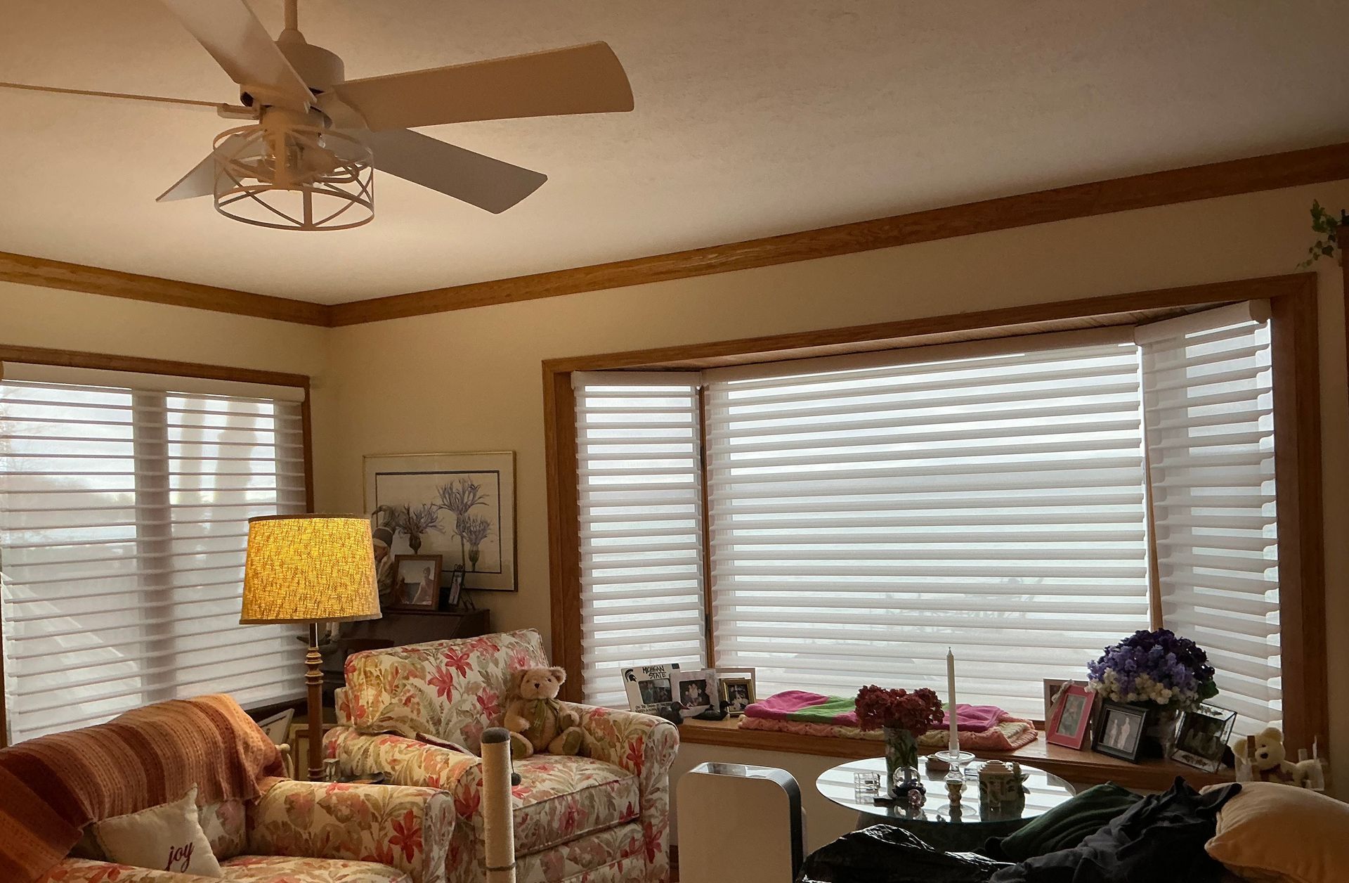 Cozy living room with floral armchairs, blinds, ceiling fan, lamp, and glass coffee table by large windows