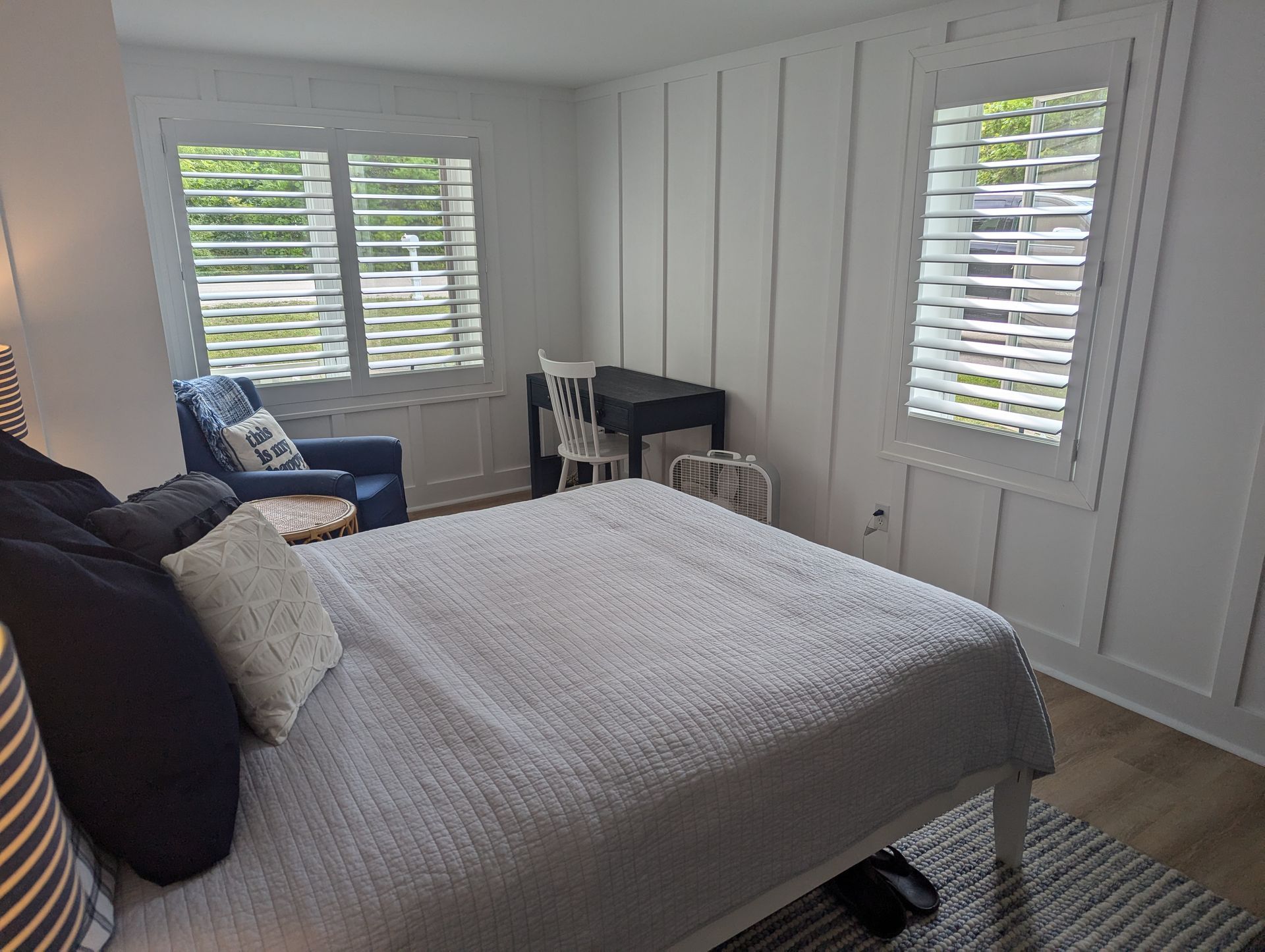 Bedroom with gray bedspread, white shutters, black chair, and a small desk by the window.
