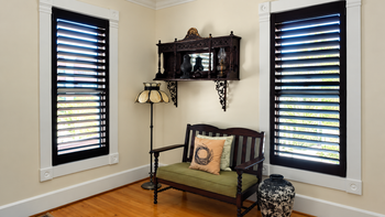 Small corner room with a black loveseat, wall shelf, lamp, and two tall windows with dark blinds.