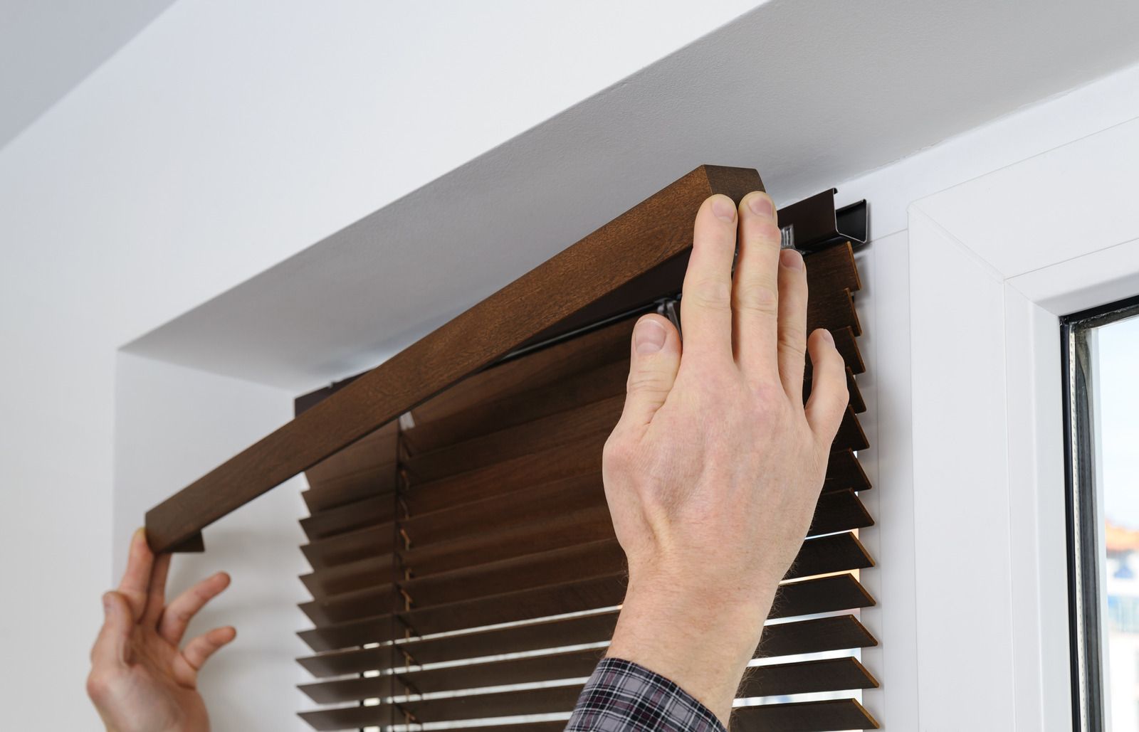 Hands adjusting a brown window blind near a white window frame