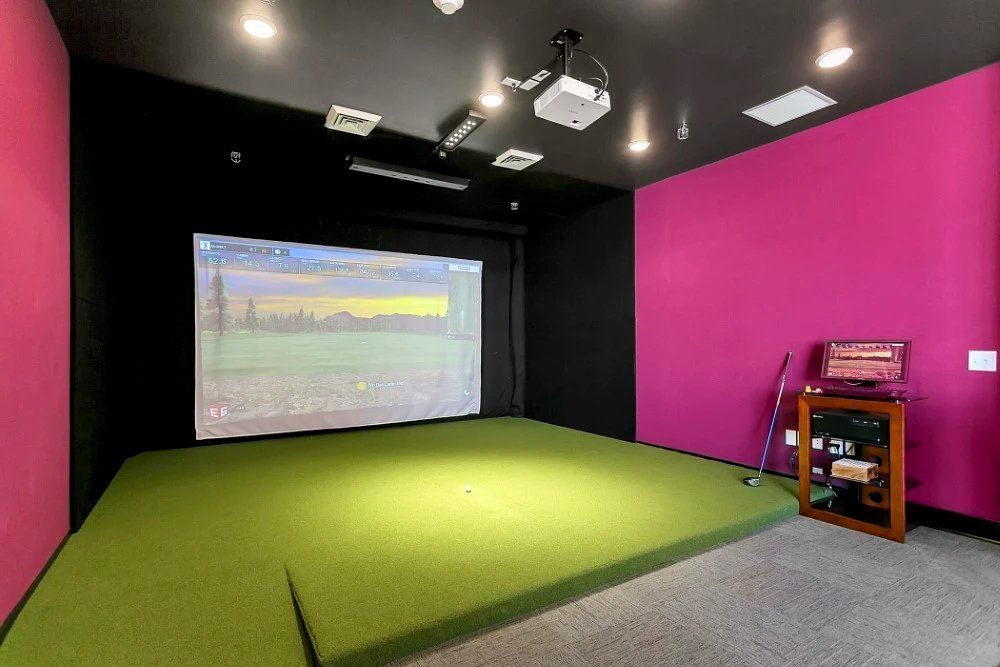 Golf simulator room with green turf, projector screen, and magenta walls.