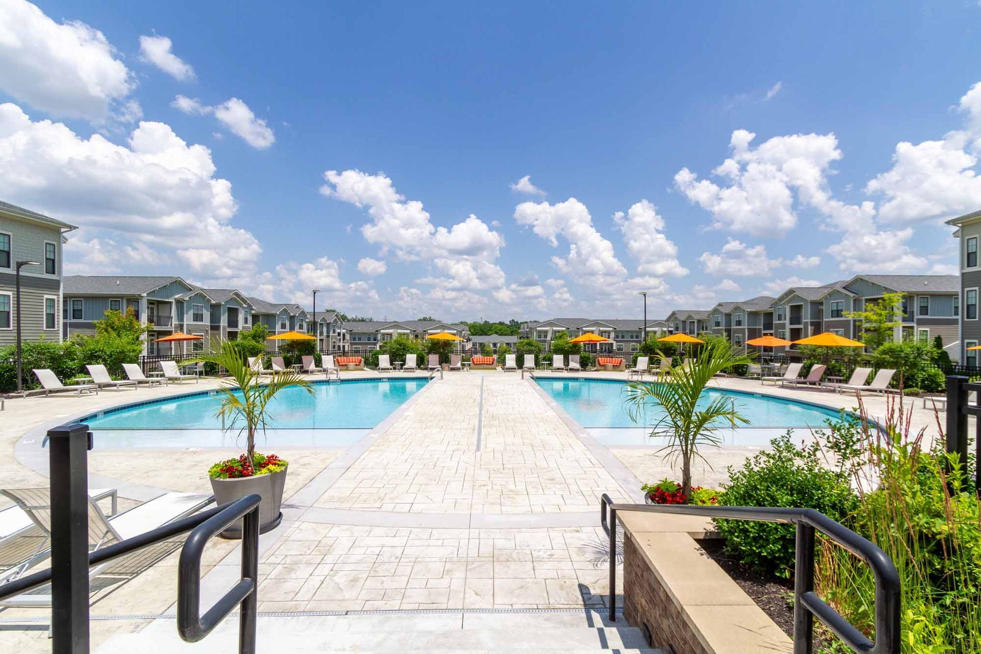 Two symmetrical pools with lounge chairs, surrounded by apartment buildings under a blue sky with clouds at Haven on Tucker Apartments in Louisville, KY