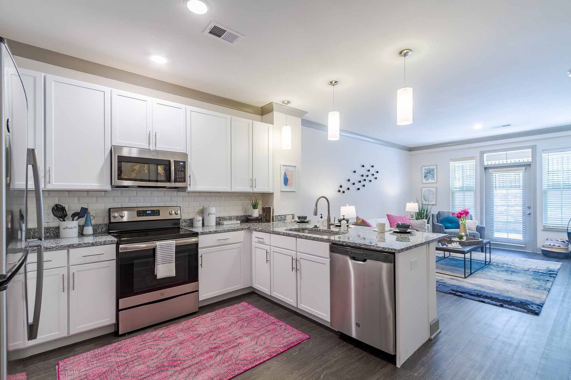 Bright, modern kitchen with white cabinets, stainless steel appliances, and a pink rug, opening to a living area at Haven on Tucker Apartments in Louisville, KY