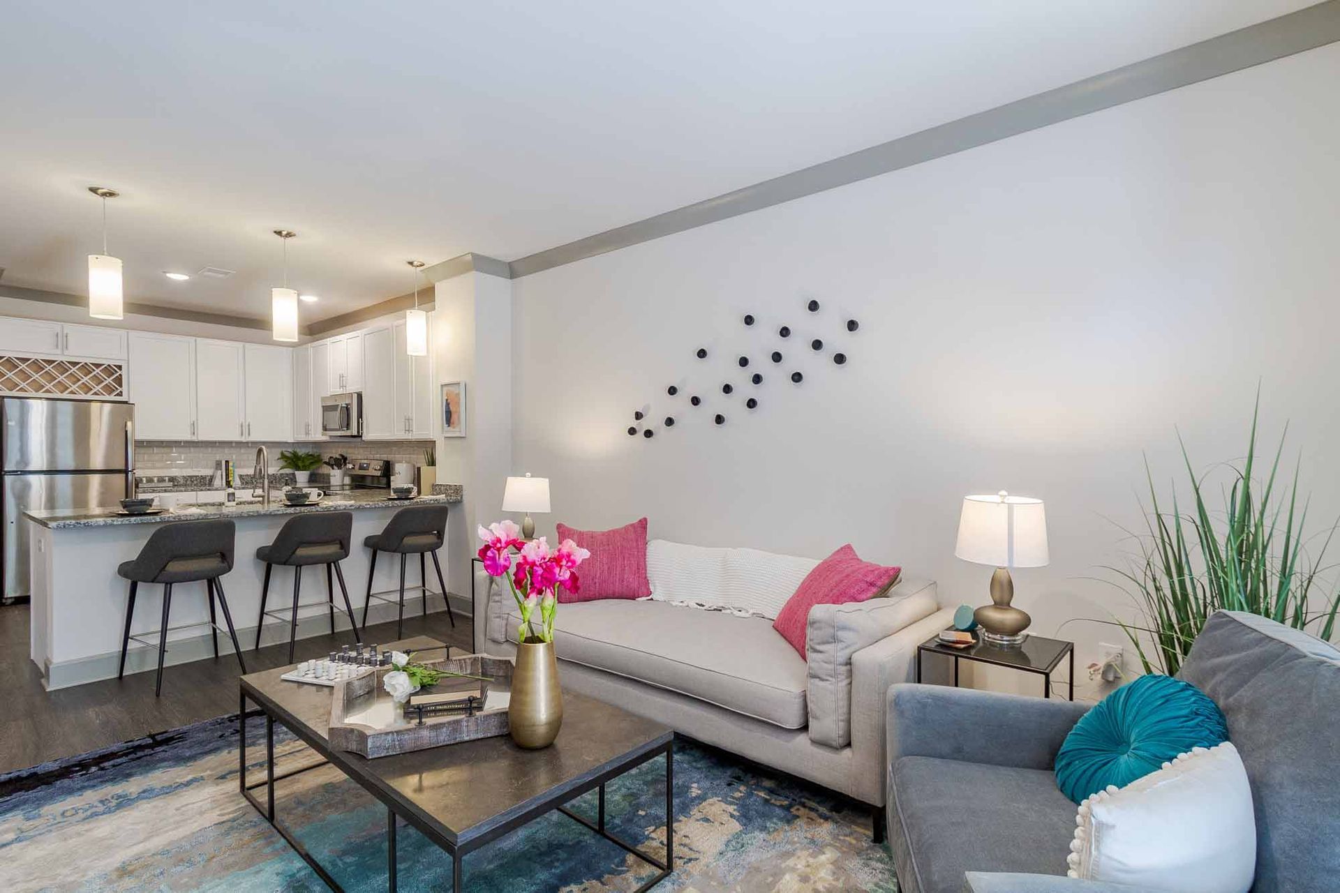 Open-concept living space with kitchen and seating area. White walls, grey trim, neutral sofa with pink pillows, and a patterned rug at Haven on Tucker Apartments in Louisville, KY