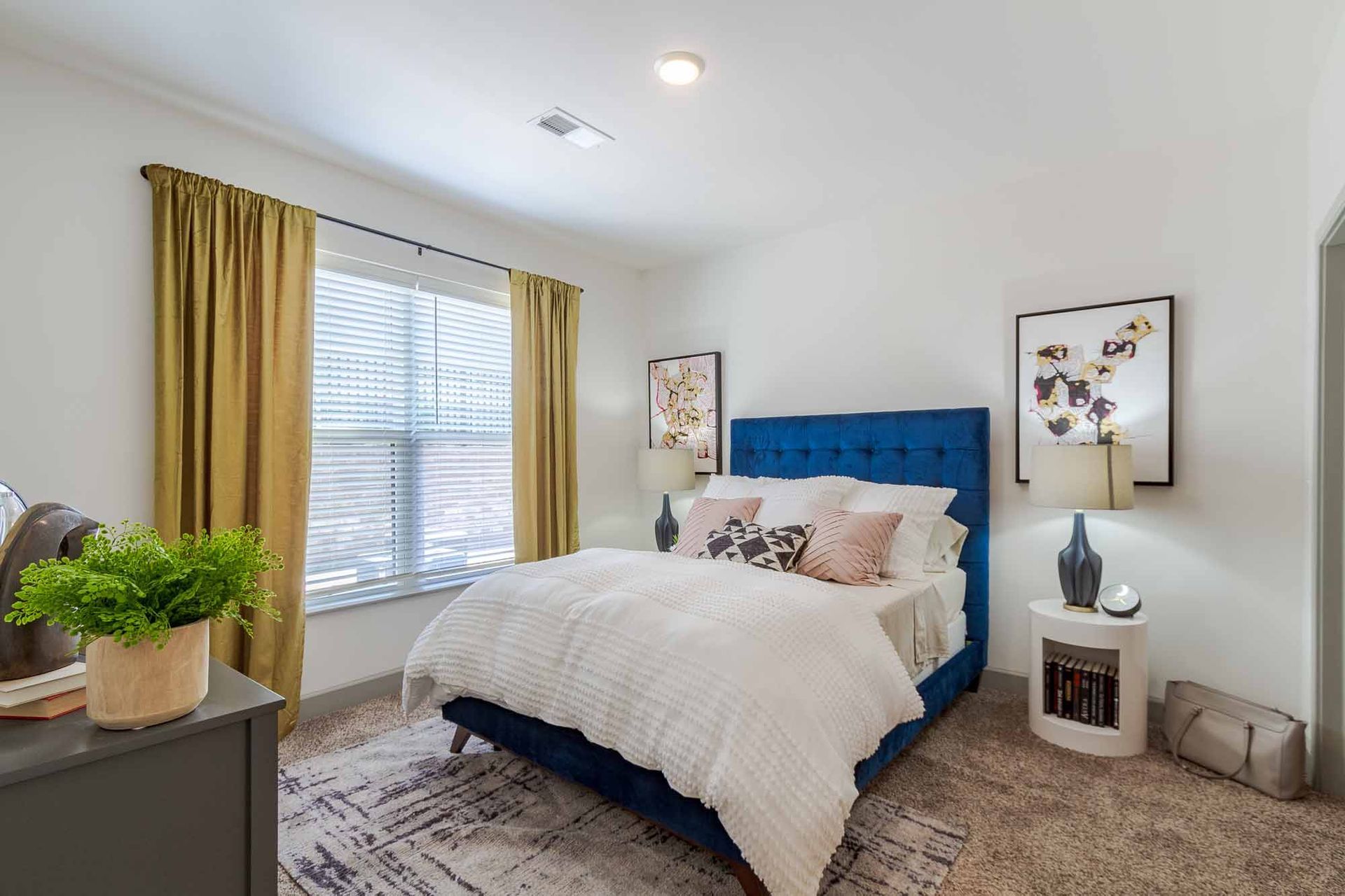 Bedroom with blue velvet bed, white bedding, gold curtains, and a plant on a gray dresser at Haven on Tucker Apartments in Louisville, KY