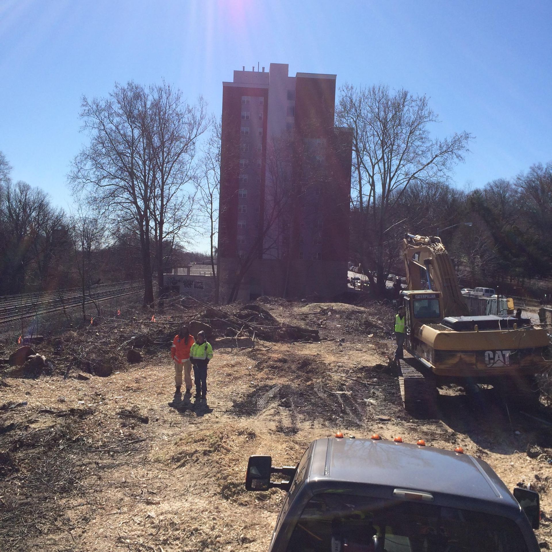 Construction site with a tall brick building, excavators, and two workers in front of bare trees under a sunny sky.