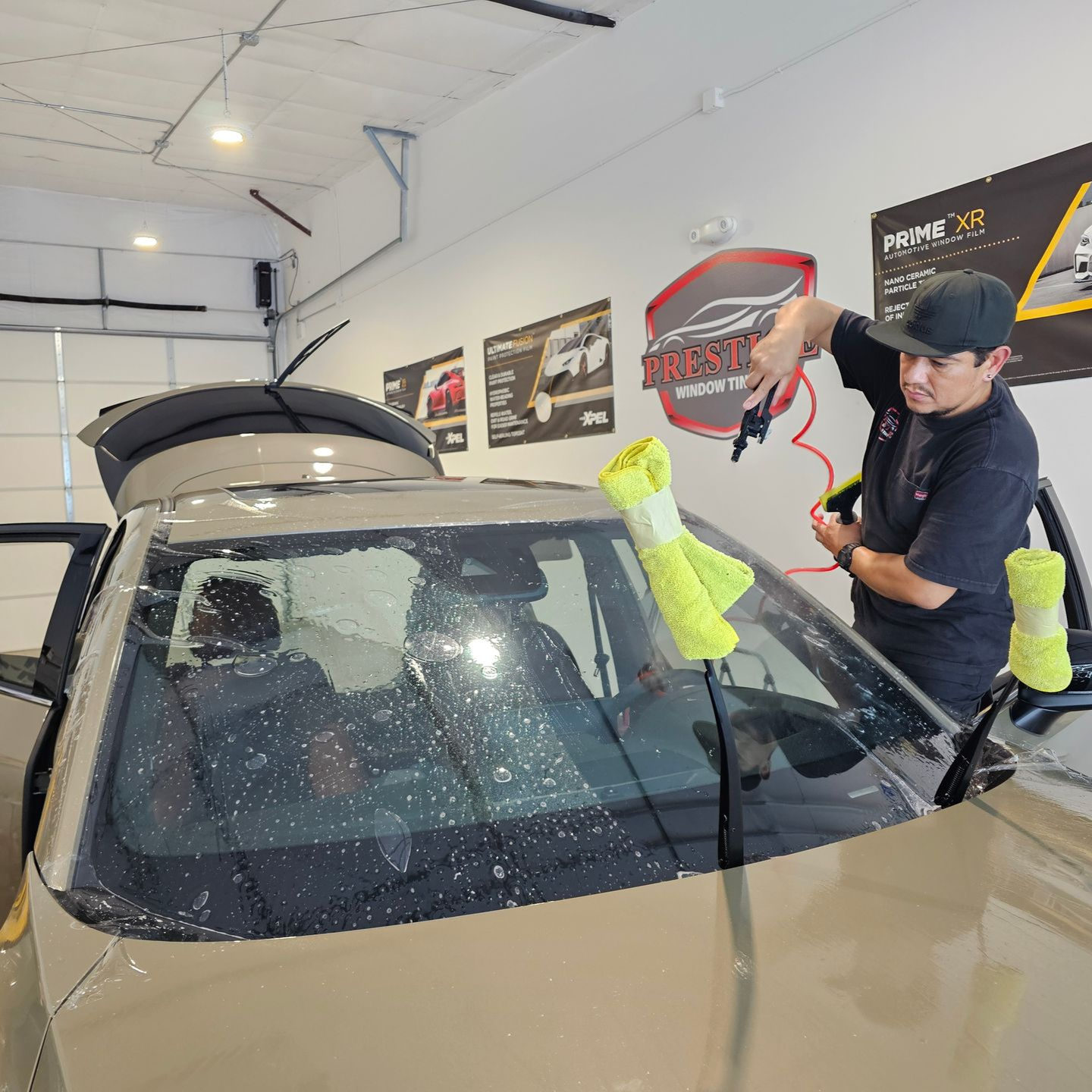 Person in a garage washing a car windshield with a red hose sprayer, water droplets on the glass.