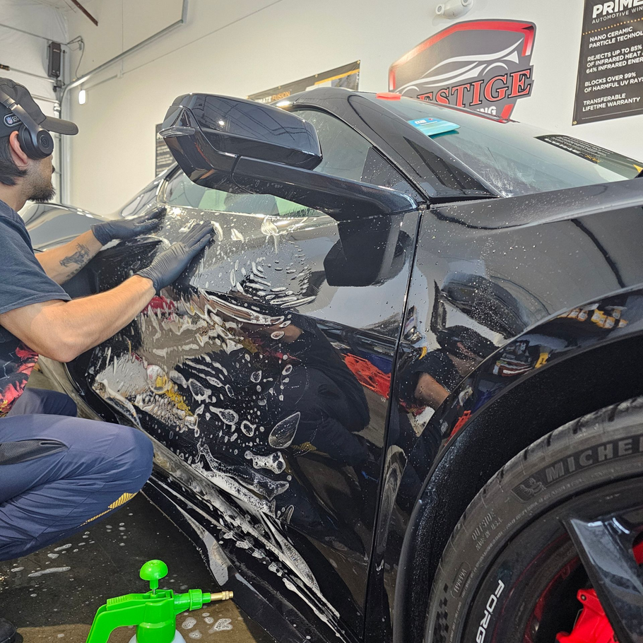 Detailer polishing a black car in a garage, with soapy water and a clay bar on the door.