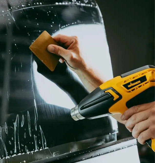 Hands using a yellow drill and sanding block on a glossy black car surface
