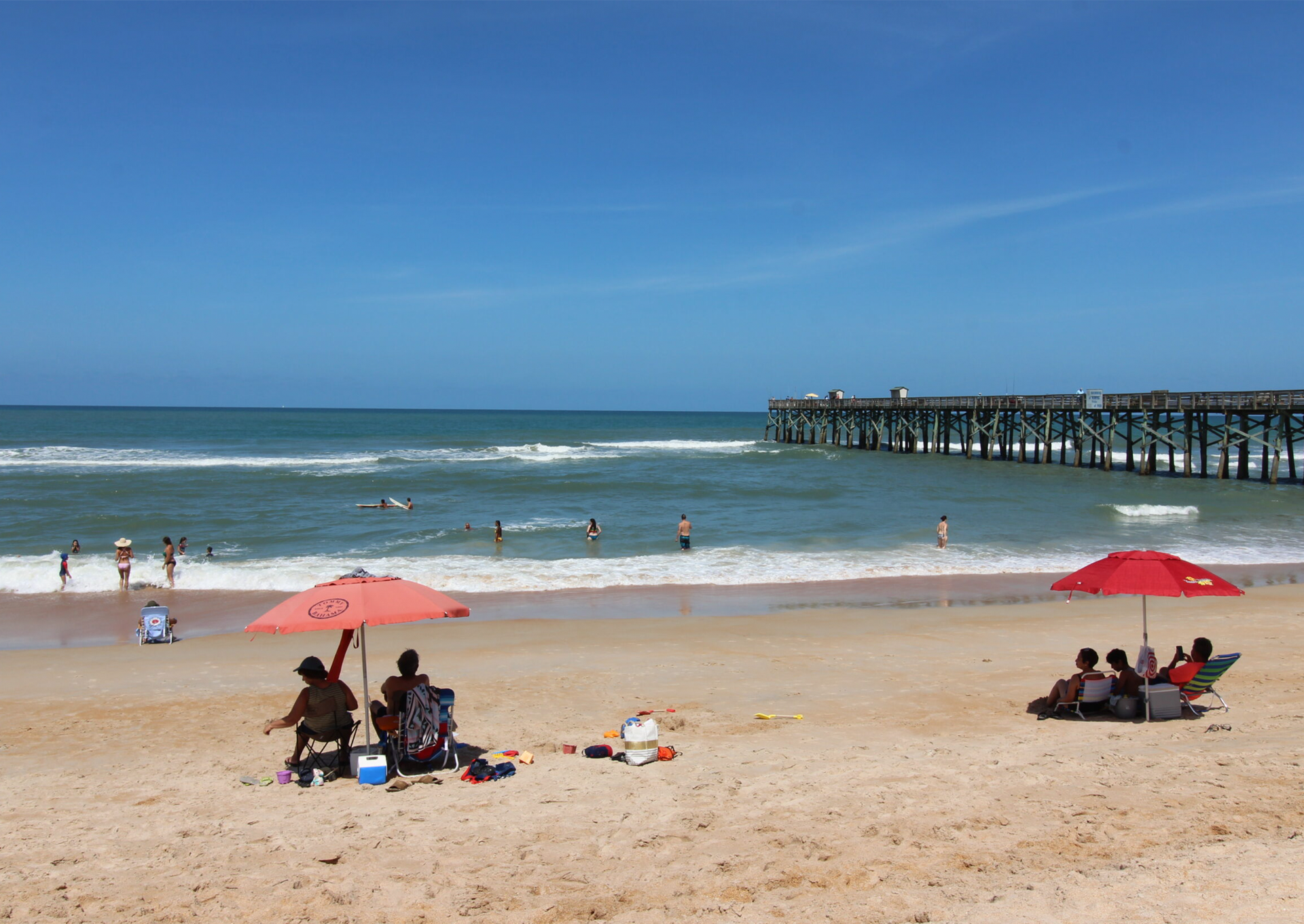 People are sitting under umbrellas on a beach near a pier