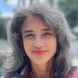 Smiling person with gray hair and pink lipstick, outdoors with a blurred sky background
