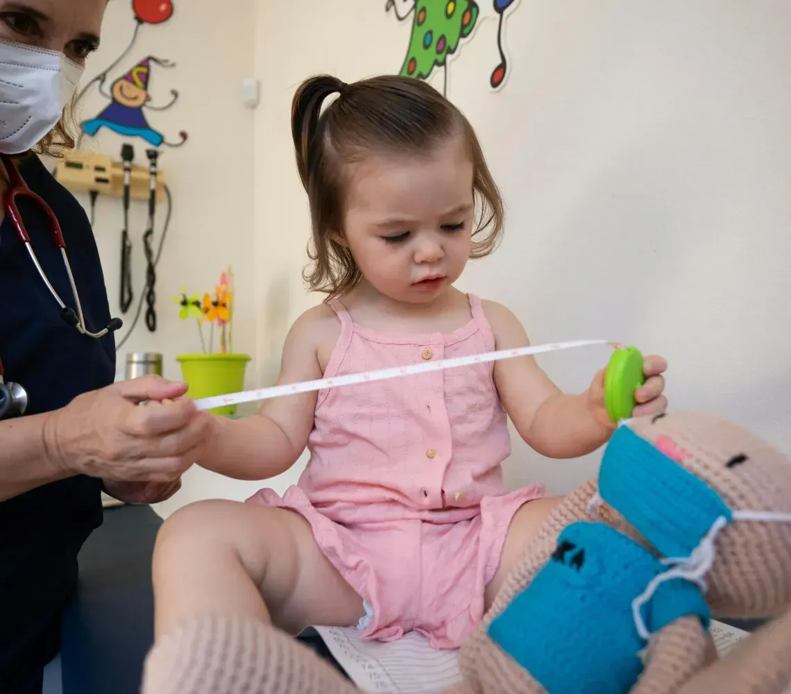 Child in pink dress sitting on a caregiver’s lap, holding a toy in a colorful clinic room.
