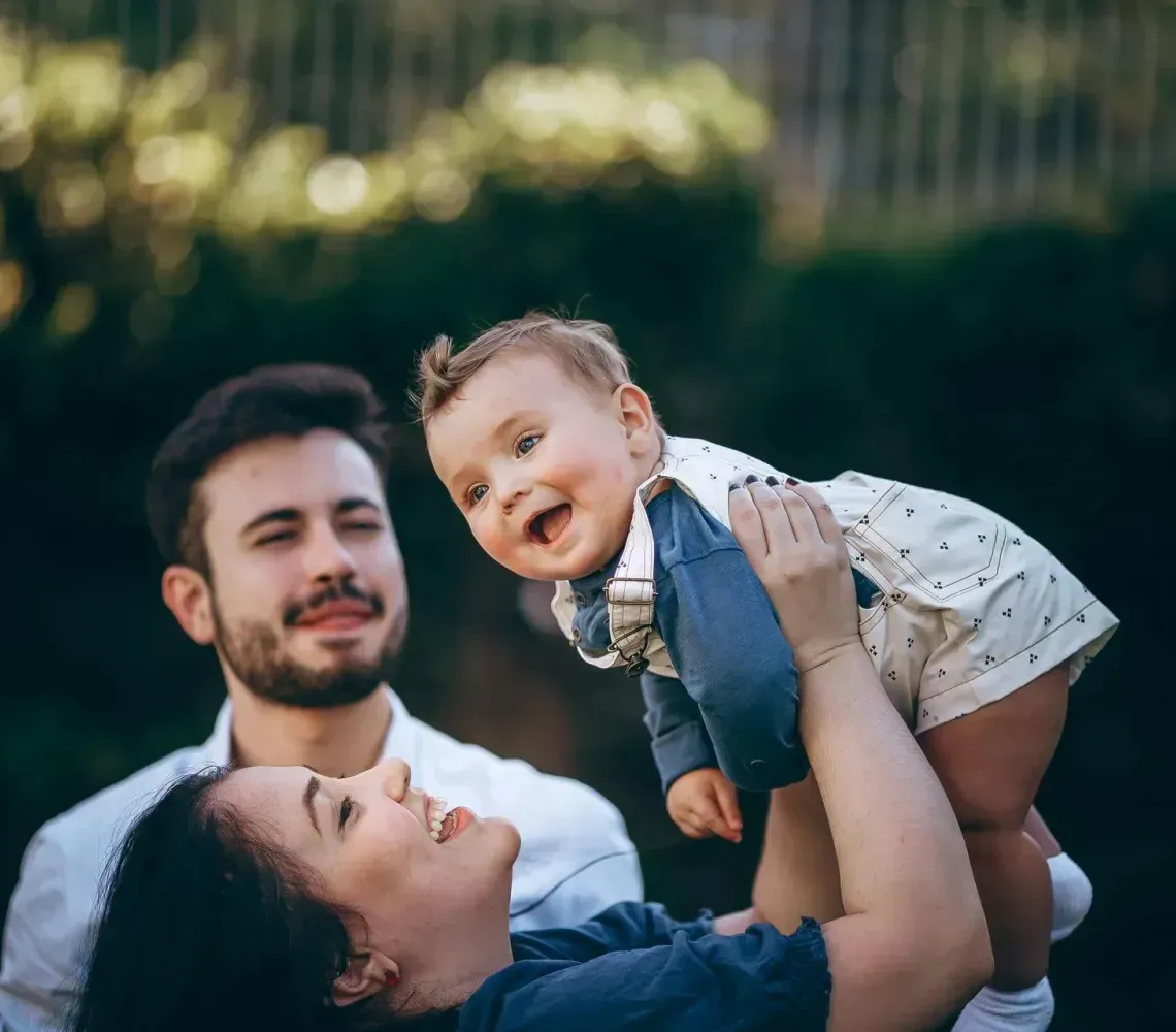 Family outdoors, lifting a smiling baby against a blurred green background.