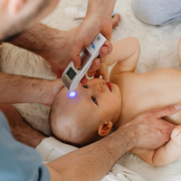Parent using a digital thermometer on a baby lying on a bed, with a glowing tip near the baby's forehead