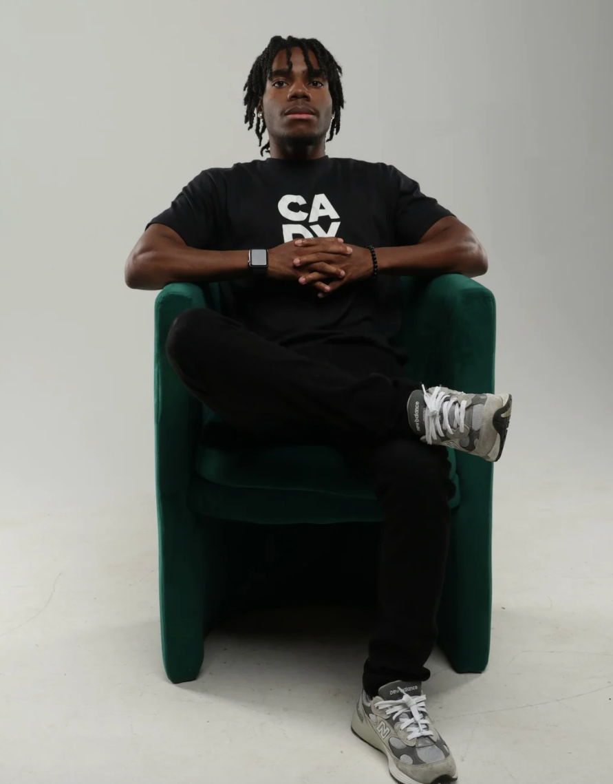 Person seated in a green chair against a plain background, wearing a black T-shirt and gray sneakers.