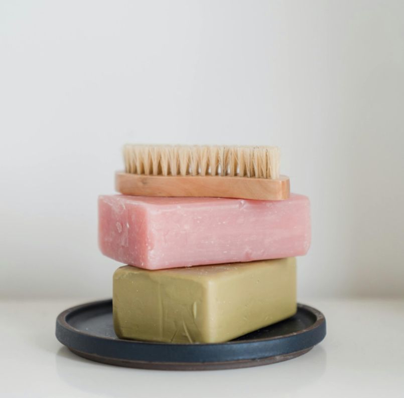 Stacked pink and olive soap bars with a wooden brush on a dark dish against a white background