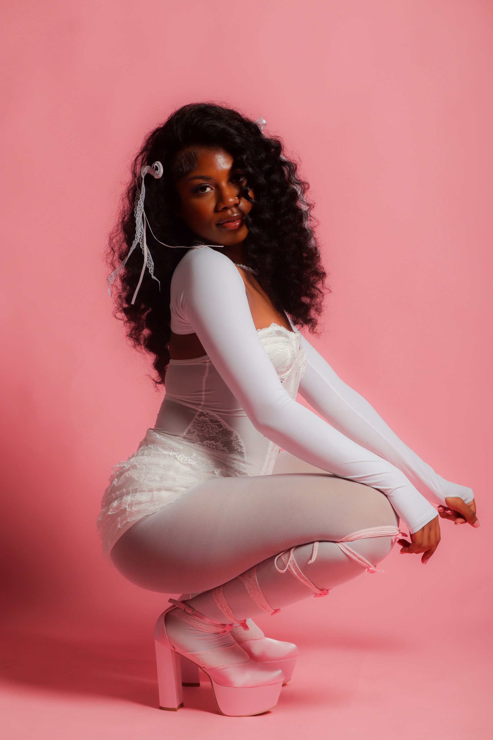 Woman crouching in a white outfit and pink heels against a pink backdrop.