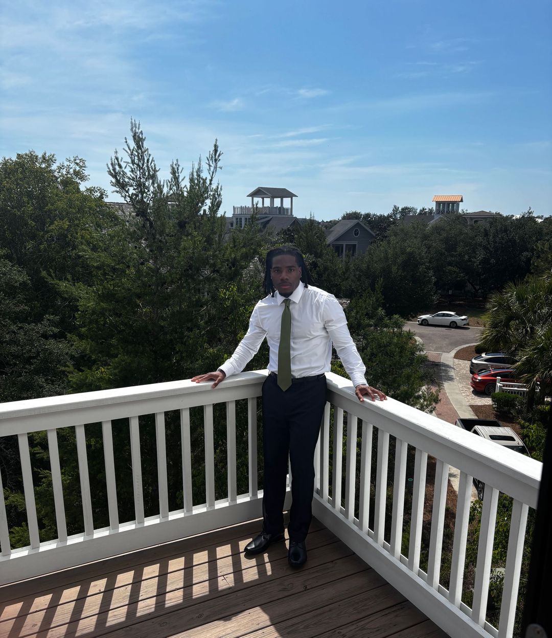 Person in white shirt and tie standing on a balcony overlooking trees and rooftops on a sunny day