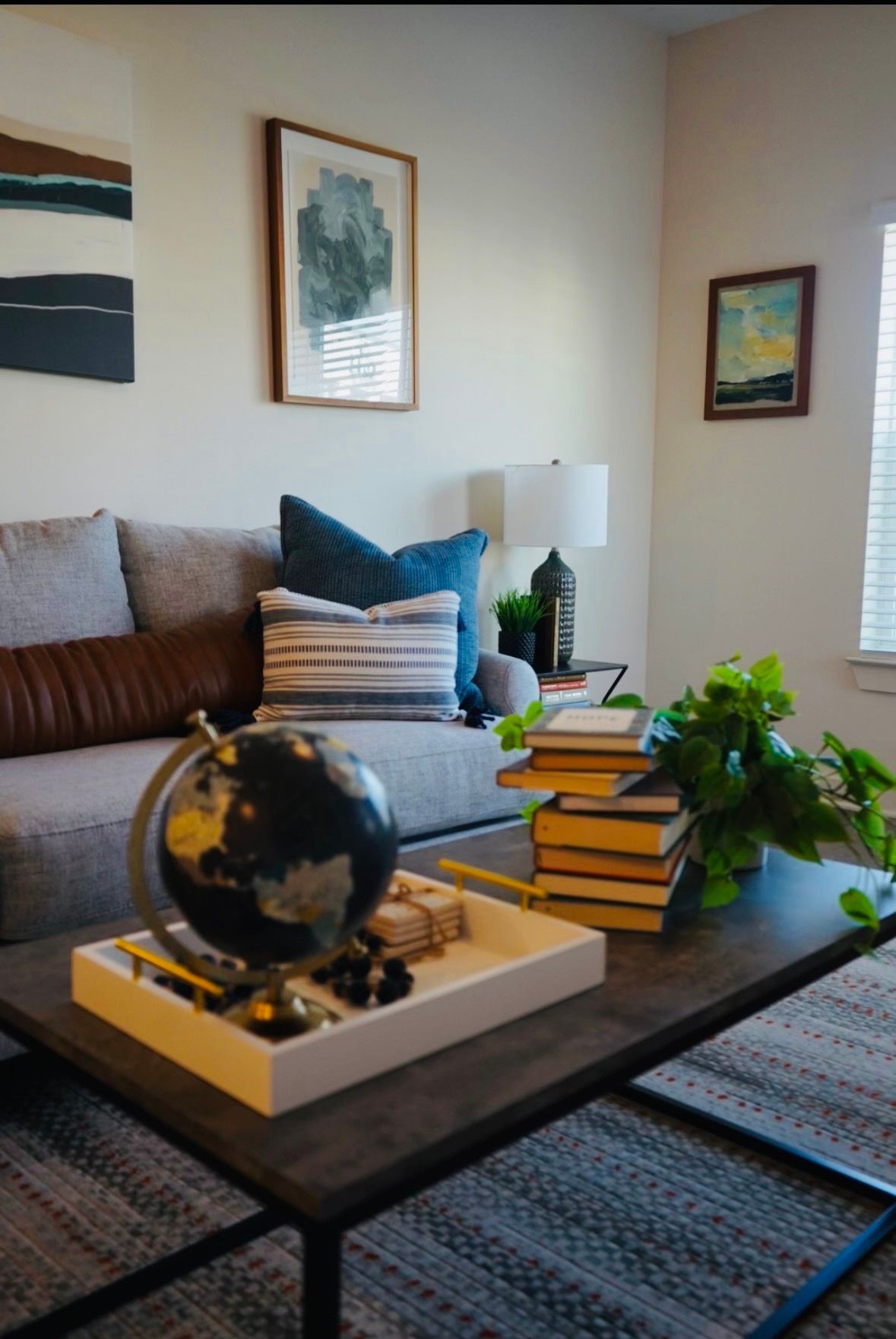 Cozy living room with gray sofa, framed art, table lamp, globe, books, and green plant on a coffee table