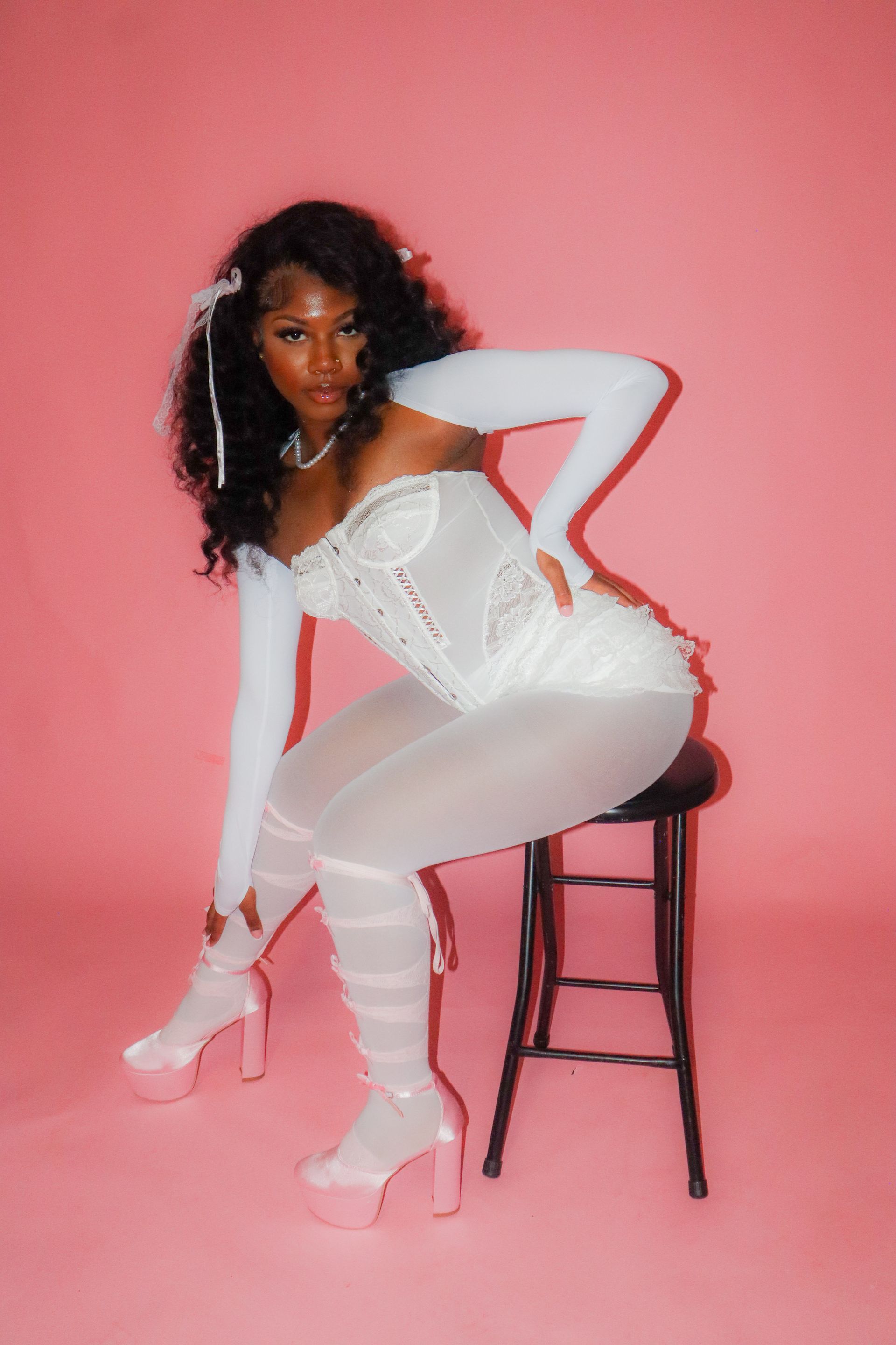 Woman posing in a white outfit and pink heels beside a stool against a pink background