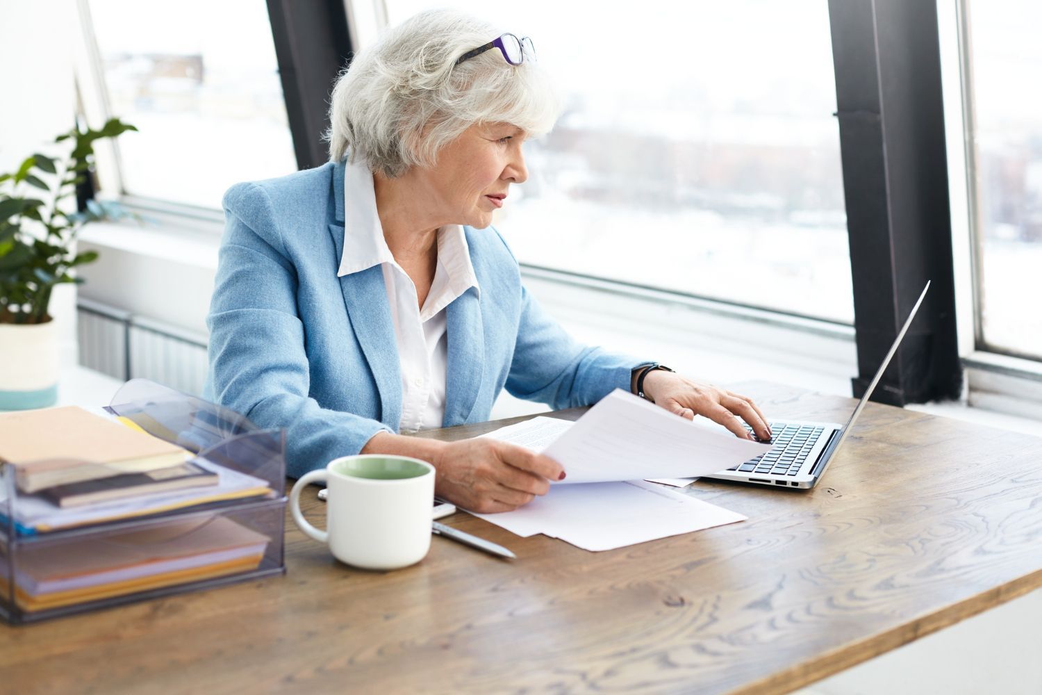 Woman in a blue blazer working on a laptop at a wooden desk by a bright window