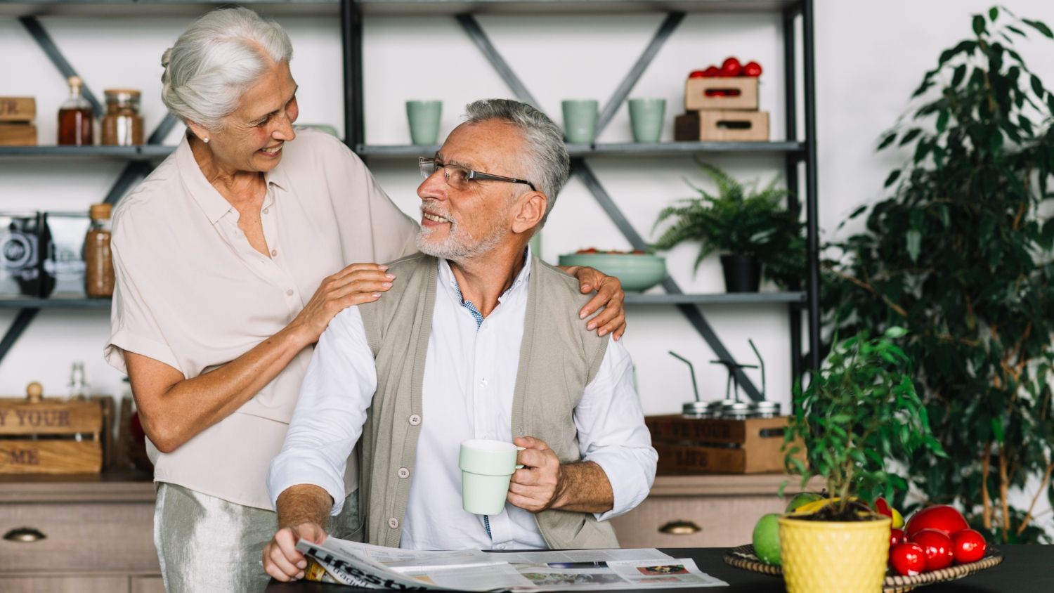 Older couple smiling in a bright kitchen, woman resting a hand on the man's shoulder as he holds a mug.