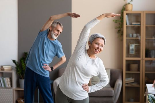 Two adults stretching in a living room, one in blue and one in white, with arms raised overhead.