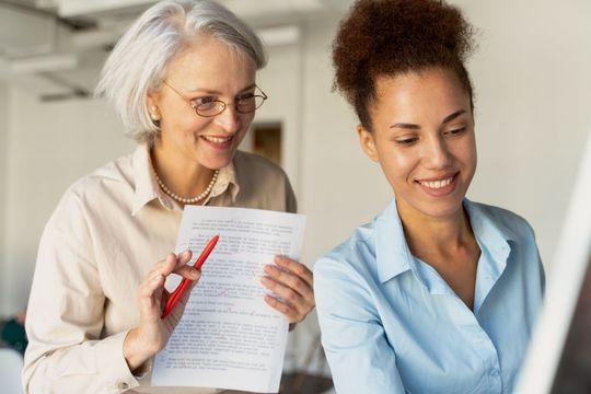 Two women reviewing a document, one pointing with a red pen in a bright office