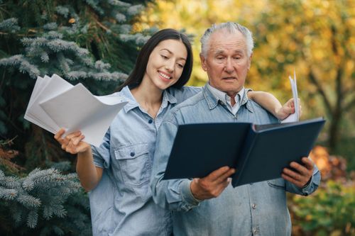 Two people outdoors reading from a large book, with papers in hand, in front of evergreen trees